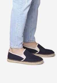 Espadrilles de malla marinas con acentos beige, detalles elásticos en los laterales y suelas de yute trenzado, combinadas con jeans de tiro vuelto en azul claro.