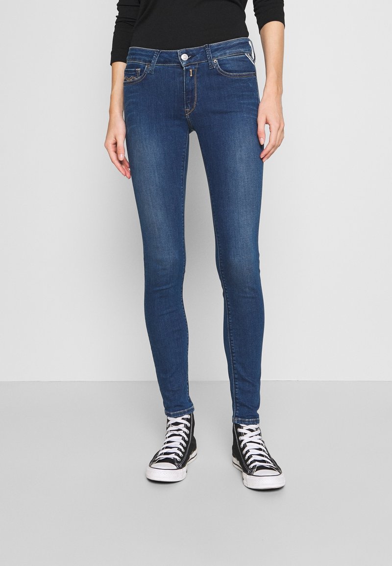 Jeans ajustados de mezclilla azul oscuro, con un diseño clásico de cinco bolsillos, detalles sutiles de costura y una cinturilla con trabillas. Combinados con zapatillas altas negras.