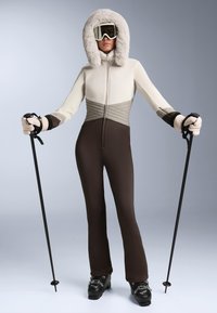 Beige és barna síoverall, szőrmével bélelt kapucnival, steppelt felsőrésszel és testre szabott kialakítással. Fekete síbakancs és síbotok egészítik ki a szettet.