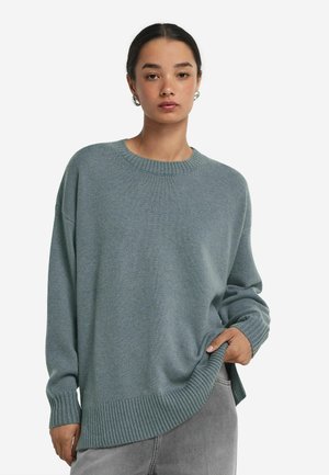 Ung kvinde iført en løs lyseblå striktrøje og grå bukser, stående med den ene hånd i lommen mod en ensfarvet hvid baggrund.
