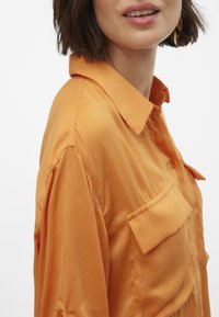Camicetta arancione con bottoni, realizzata in tessuto liscio, dotata di colletto punta, taschino sul petto e vestibilità rilassata con maniche arrotolate.