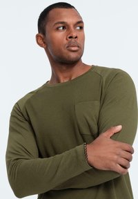 Camicia a maniche lunghe di colore verde oliva realizzata in tessuto a coste, con colletto girocollare e tasca sul petto. Il tessuto ha una finitura liscia e le maniche sono aderenti.