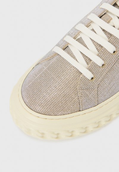 Sneaker en tissu métallique doré clair avec des accents beige, présentant une surface texturée, un bout rond et des lacets crème. Semelle en caoutchouc avec un bord décoratif.