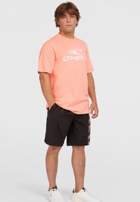 Camiseta de color rosa claro con el logo blanco de "O'Neill", combinada con pantalones cortos negros con un panel lateral estampado y zapatillas blancas.