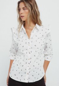 Witte overhemd met knoopjes en een klein bloemenpatroon, met ruches op de schouders en opgerolde mouwen. Het materiaal lijkt licht van gewicht te zijn.