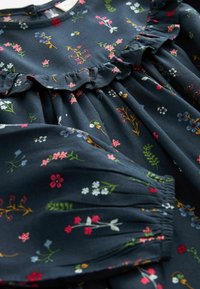 Tissu bleu foncé avec de petits imprimés floraux multicolores, avec une couture froufroutante au niveau de la poitrine et des poignets élastiqués froncés aux manches.
