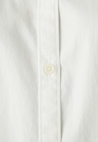 Biała koszula z kołnierzykiem, z detalem guzików, charakteryzująca się gładką, jednolitą teksturą oraz widocznym przeszyciem wzdłuż plisy.