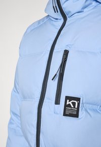 Giacca puffa azzurro chiaro con colletto alto, tasche con zip nere e una patch del logo del marchio nera sul davanti. Texture liscia e trapuntata.