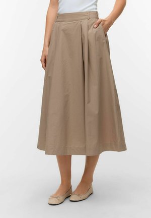 Femme portant une jupe midi beige à plis avec des poches, associée à des ballerines beiges et un haut blanc, debout sur un fond uni.