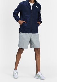 Man wearing navy zip-up jacket, light gray shorts, and white athletic sneakers standing with hands in pockets.