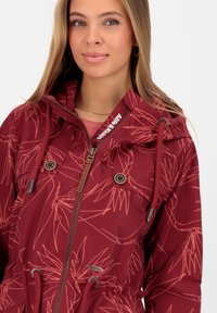 Giacca bordeaux con motivo floreale, cappuccio con cordino, zip frontale e tasche a scatto. Tessuto testurizzato con un design a vita cinchata.