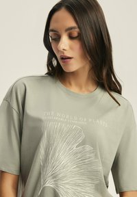 Zielona bawełniana koszulka z krótkim rękawem, prezentująca biały grafikę liścia oraz napis "ŚWIAT ROŚLIN NIEWZRUCHOMA SYMBIOZA". Okrągły dekolt.