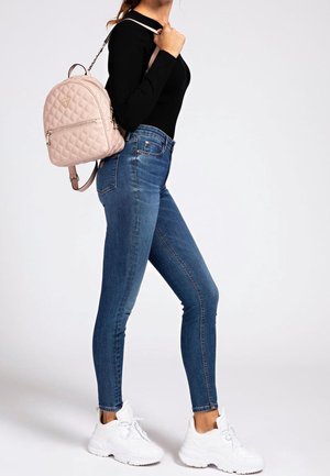 Frau trägt schwarzes Oberteil, blaue Skinny-Jeans, weiße Sneakers und hat einen hellrosa gesteppten Rucksack über einer Schulter getragen.