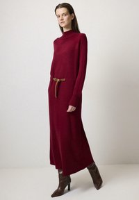 Rochie tricotată burgundă cu mâneci lungi și guler înalt, încinsă în talie cu o curea din piele maro, purtată cu bocanci maro cu toc.