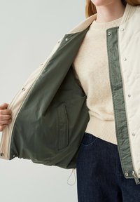 Gepolsterte Oberbekleidung mit einer cremefarbenen Außenseite und grüfem Futter, gesichert durch silberne Hardware. Darunter befindet sich ein weicher gestrickter Creme-Pullover.