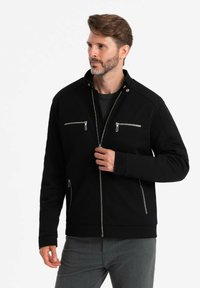 Veste noire en tissu texturé, avec un col montant, des poches zippées et une fermeture éclair à l'avant. Ajustement grande taille avec des manches s'étendant jusqu'aux poignets.