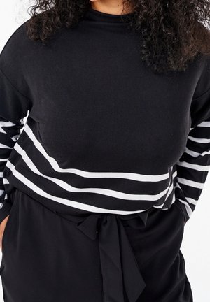 Femme portant un haut noir à manches longues avec des bandes blanches sur les manches et près de la taille, associé à un pantalon noir à taille nouée.