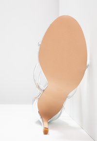Sandale transparente avec une semelle beige et un talon fin, positionnée verticalement contre un angle blanc entre le mur et le sol.