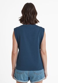 Person with shoulder-length brown hair wearing a sleeveless dark blue top and rolled-up denim shorts, facing away against a white background.