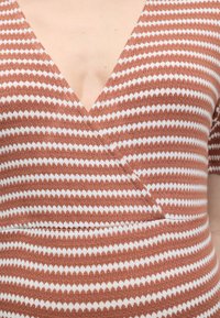 Καφέ και λευκό πλεκτό ρούχο με μοτίβο ζιγκ-ζαγκ και V-λαιμόκοψη, φορεμένο στο πάνω μέρος του κορμού με κοντά μανίκια.