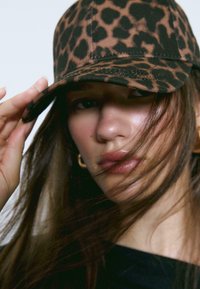 Brun og sort leopardprint baseballcap med en buet skygge, lavet af stof, med syet detalje langs kanten af skyggen.