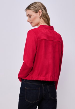 Femme aux cheveux blonds vêtue d'une veste rouge et d'un jean bleu foncé, légèrement tournée de profil sur un fond clair uni.