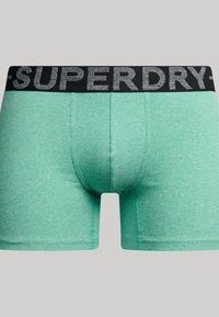 Mintgroene boxershorts met een zwarte elastische tailleband waarop "SUPERDRY" in grijze tekst staat, gemaakt van een zachte, gestructureerde stof.