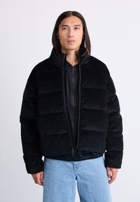 Giacca puffer in corduroy nera con colletto alto, chiusura con zip e motivo trapuntato orizzontale. Indossata sopra una maglia nera con zip.