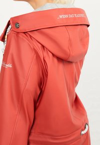 Red waterproof jacket with a drawstring hood, textured finish, white hardware accents, and printed text on the collar.