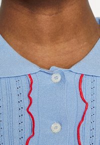 Light blue knitted polo collar with red wavy accents, featuring two button closures and textured patterns along the fabric.