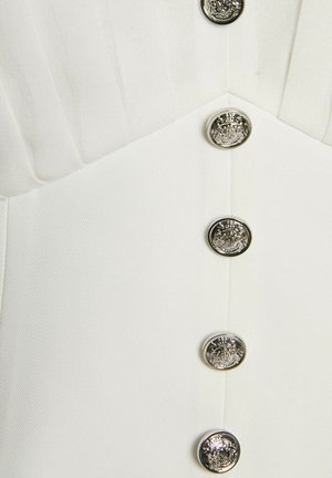 Primo piano di un indumento bianco con pieghe verticali e cinque bottoni rotondi metallici con rilievi di stemmi ornati.