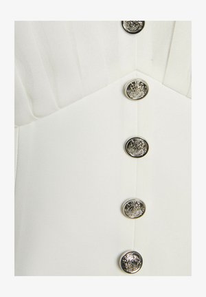 Primo piano di un indumento bianco con pieghe verticali e cinque bottoni rotondi metallici con rilievi di stemmi ornati.