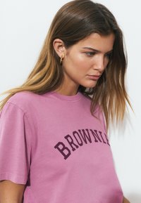 Mujer joven con cabello largo castaño, usando aretes de aro dorados y una camiseta rosa con el texto "BROWNIE", mirando hacia abajo sobre un fondo blanco.