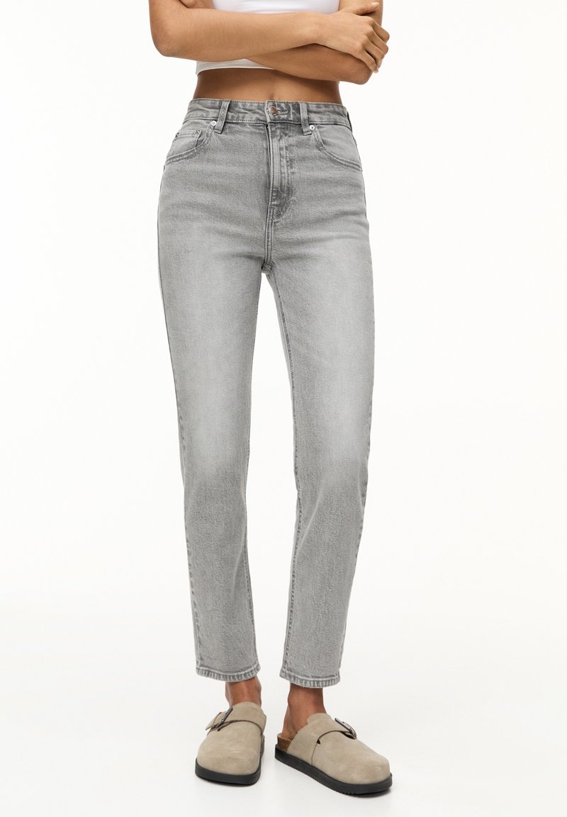 Femme portant un jean taille haute gris clair, les bras croisés, et des chaussures beige à enfiler avec des boucles sur fond blanc.