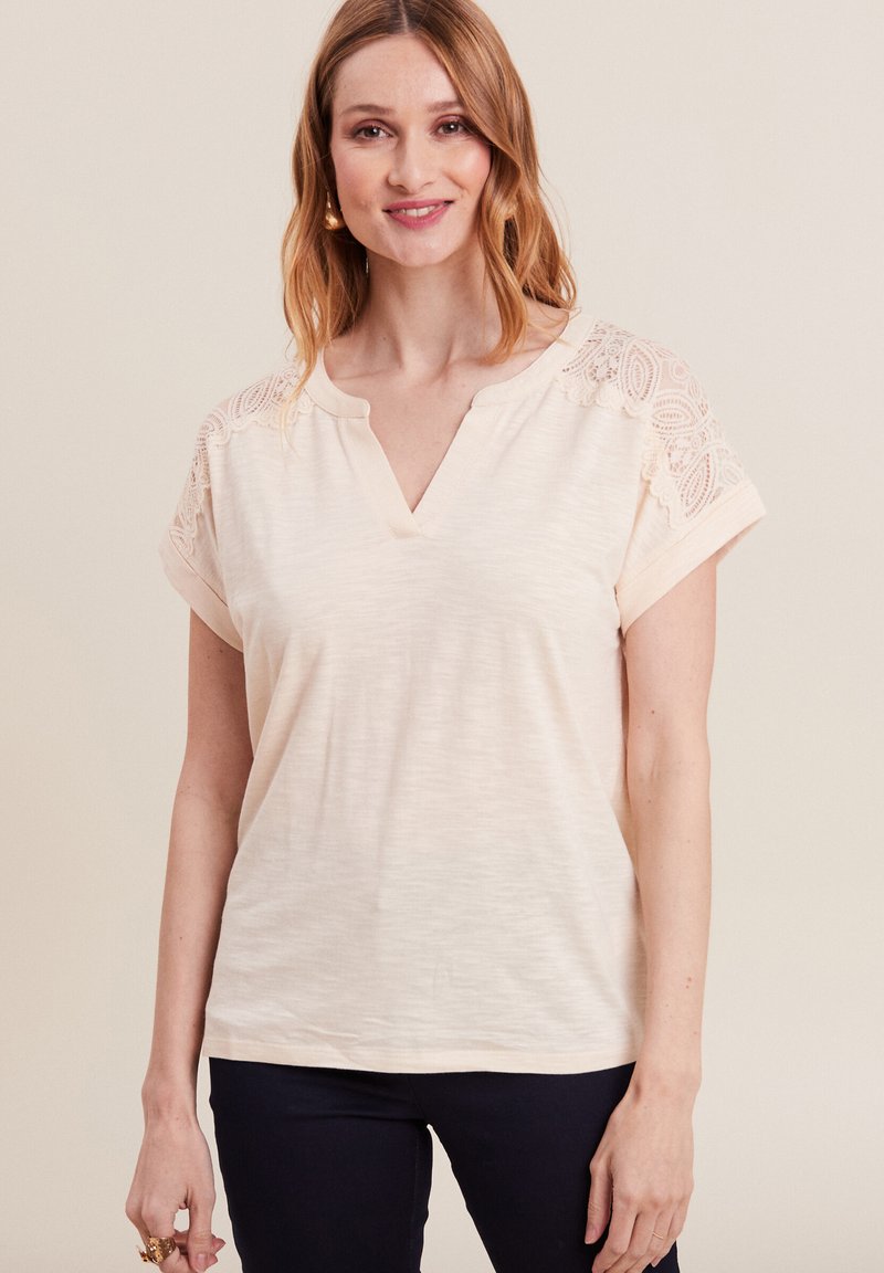 Helle beige V-Ausschnitt-Bluse mit kurzen Ärmeln, die mit Spitzenapplikationen an den Schultern versehen ist und eine lockere Passform bietet. Weicher Stoff und leichtes Material.