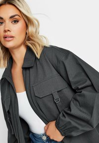 Veste bomber grise avec une fermeture éclair, coupe oversize, deux poches avant, poignets élastiques et tissu texturé. Portée par-dessus un haut blanc.