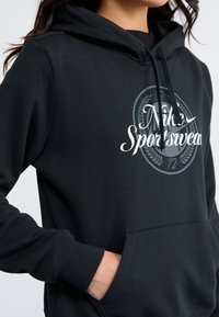 Sweatshirt com capuz preto e bolso frontal, com gráficos brancos e o logótipo "Nike Sportswear" no peito. Material em mistura de algodão.