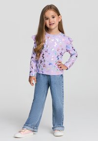 Długi rękaw fioletowej bluzki z falbanami na ramionach, z wielokolorowym wzorem gwiazd, w połączeniu z jasnoniebieskimi dżinsami i pastelowymi sneakersami.