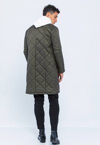 Olivgrüner gesteppter langer Mantel mit einer weißen Kapuze, versehen mit einem Rauten-Design, langen Ärmeln und geradem Schnitt. Kombiniert mit schwarzen Hosen und Stiefeln.