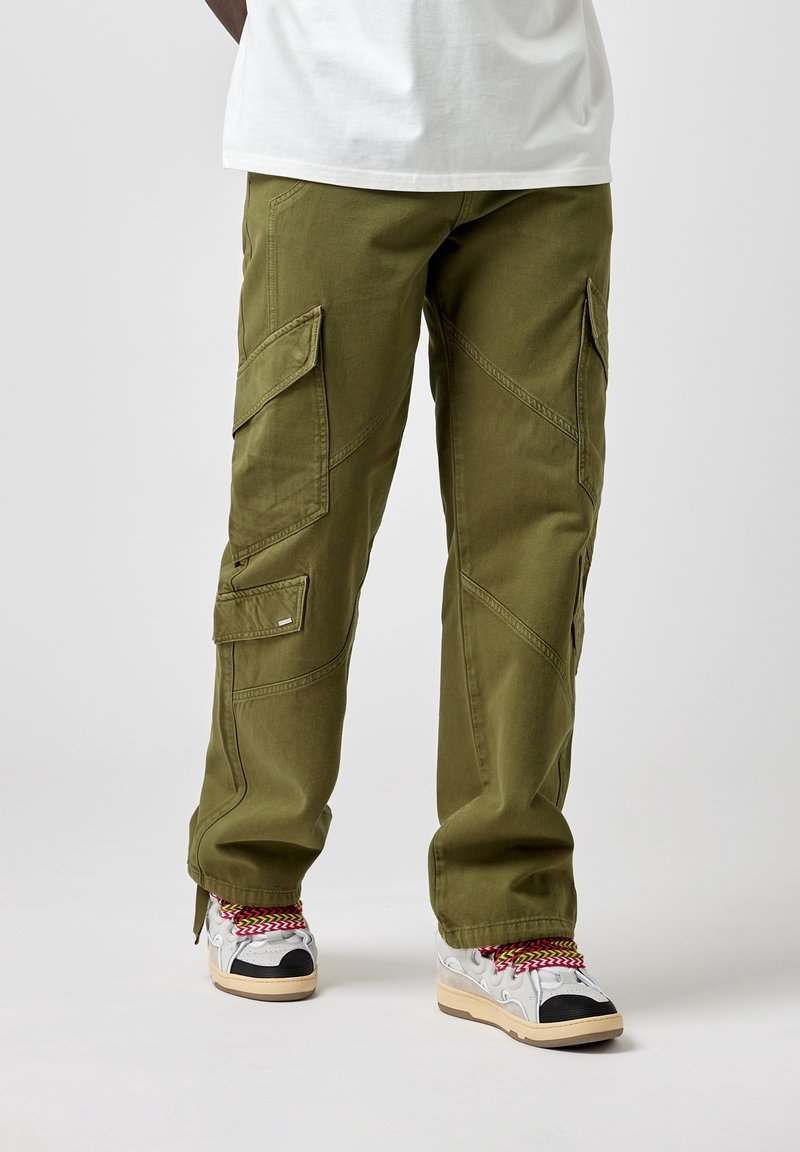 Pantalon cargo vert olive avec multiples poches, coupe fuselée et coutures en biais, associé à des baskets multicolores avec un design texturé.