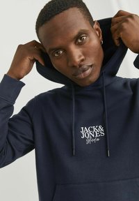 Tengeri kék kapucnis pulóver, pamutból készült, kenguru zsebbel, húzózsinóros kapucnival és fehér hímzett "JACK & JONES Studio" logóval a mellkason.