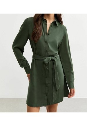 Frau trägt ein langärmeliges, dunkelgrünes Hemdblusenkleid mit einem gebundenen Stoffgürtel in der Taille, steht vor einem einfarbigen Hintergrund.