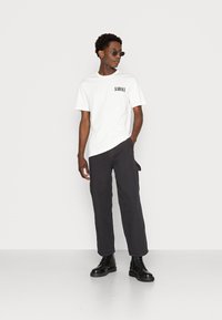 White t-shirt with "SCARFACE" print on the chest, paired with black, loose-fitting pants and black lace-up boots. Round, dark sunglasses.