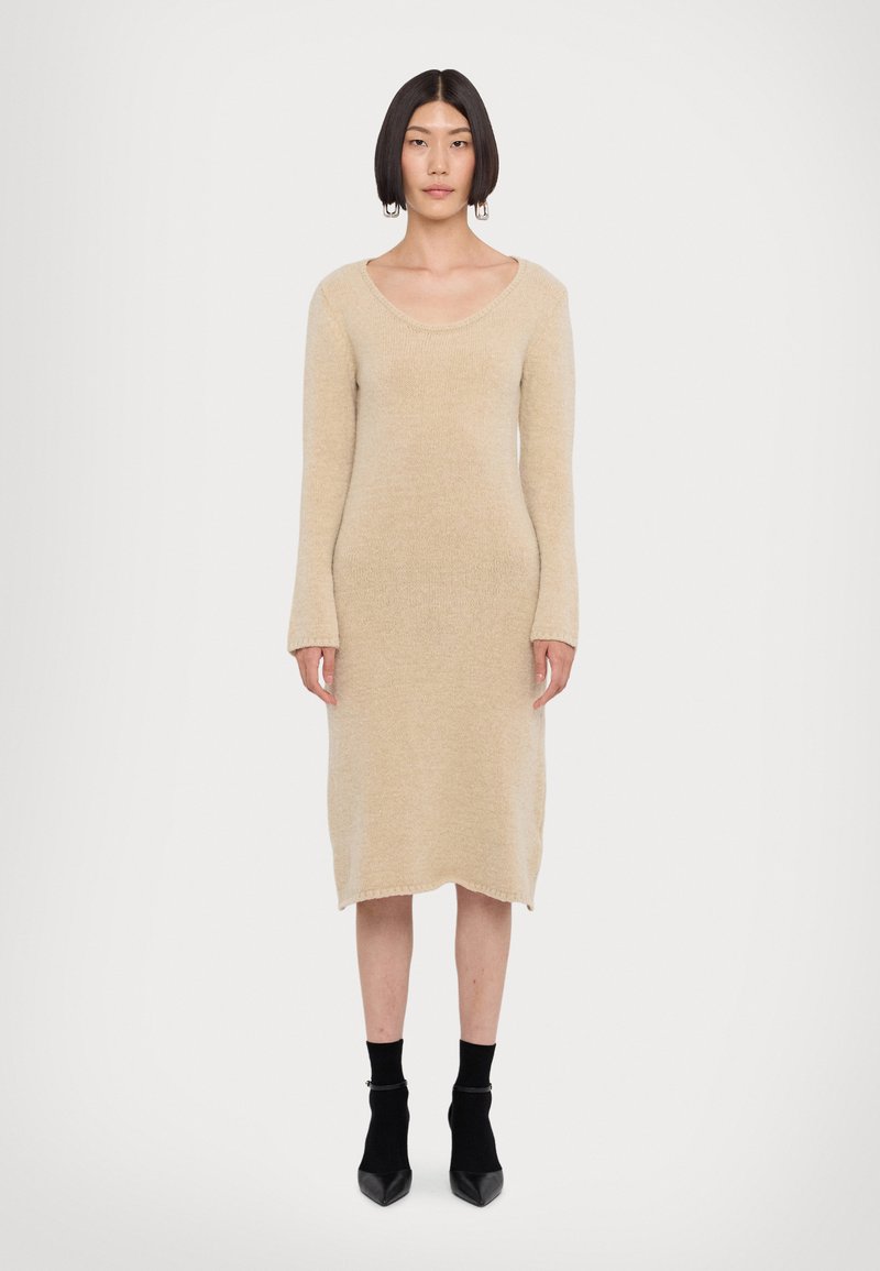 Abito maglione in beige, con scollatura a barca, maniche lunghe e orlo leggermente svasato. Indossato con calzini neri alla caviglia e scarpe a punta.