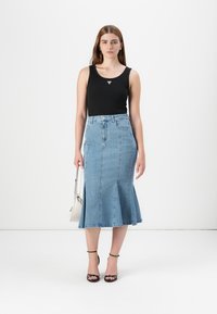 Een jonge vrouw staat zelfverzekerd, gekleed in een zwarte tanktop en een uitlopende denimrok, terwijl ze een kleine handtas vasthoudt tegen een effen achtergrond.