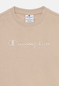 Sweat-shirt en coton beige avec un col rond, arborant un logo "Champion" brodé en blanc sur le devant et des poignets et un ourlet côtelés.