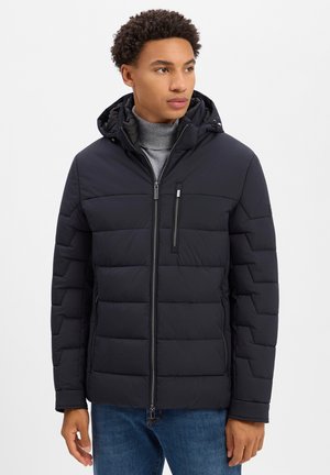 Young man wearing a black quilted hooded jacket over a gray turtleneck and blue jeans, looking slightly to the right.