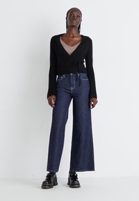 Svart strikket omslagscardigan med innsnørt midje, kombinert med høye mørke denim vide jeans. Modellen har på seg svarte platåsko.