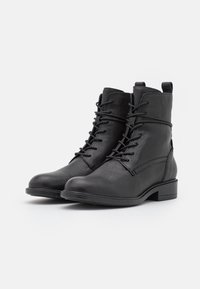 Botas de cuero negras hasta el tobillo con cordones, puntera redondeada y taco bajo. Presentan un diseño con costuras y una lengüeta en la parte trasera. Textura suave.