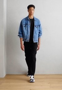 Τζάκετ τζιν, μαύρη μπλούζα, μαύρο παντελόνι και πολύχρωμα αθλητικά παπούτσια. Μοντέλο περπατάει σε ξύλινο δάπεδο μπροστά σε λευκό τοίχο.